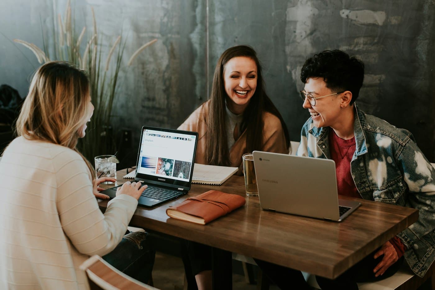 Three colleagues laughing together over laptops at a wooden table