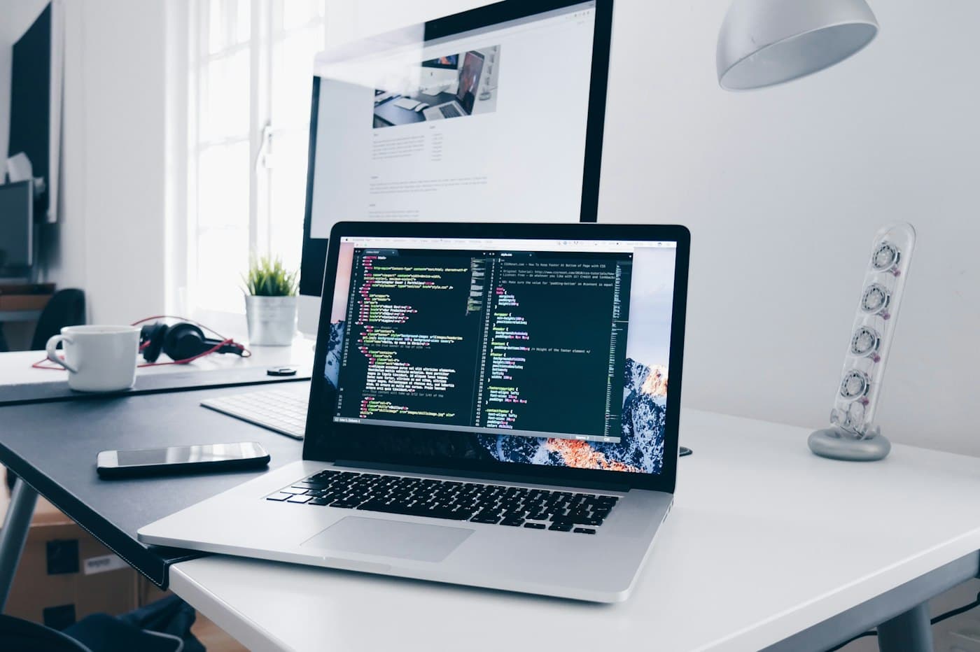Laptop showing code on a bright desk next to a monitor